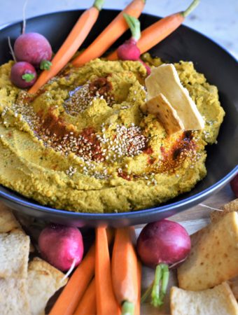 black round ceramic bowl with Indian spiced carrot hummus on a cutting board with pita chips and veggies