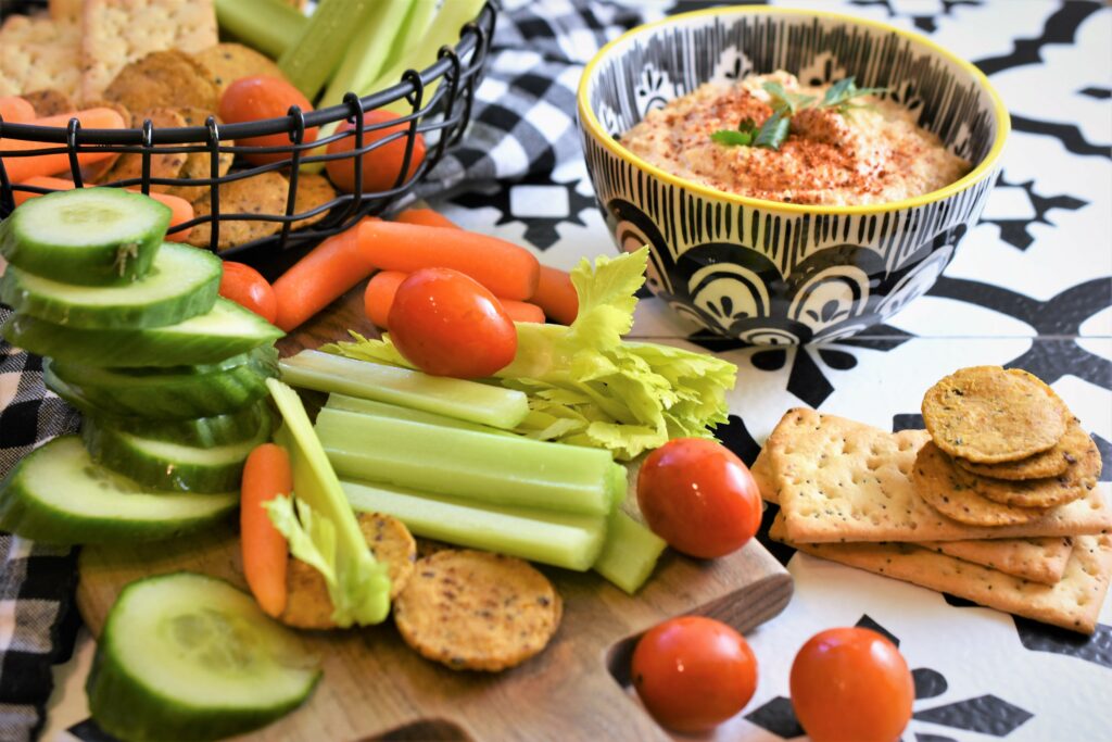 Chipotle almond dip in black and white patterned dip bowl with veggies in black wire basket off to the side with more vegetables and crackers resting on cutting board on black and white patterned tile counter top