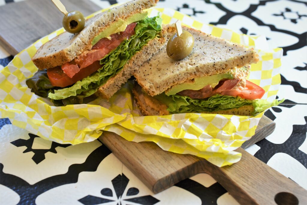 cutting board with veggies sandwich with dip used as a spread placed on top of yellow and white checked deli paper on top of handled wood cutting board on top of black and white patterned tile counter top like Joanna Gaines would use in one of her homes