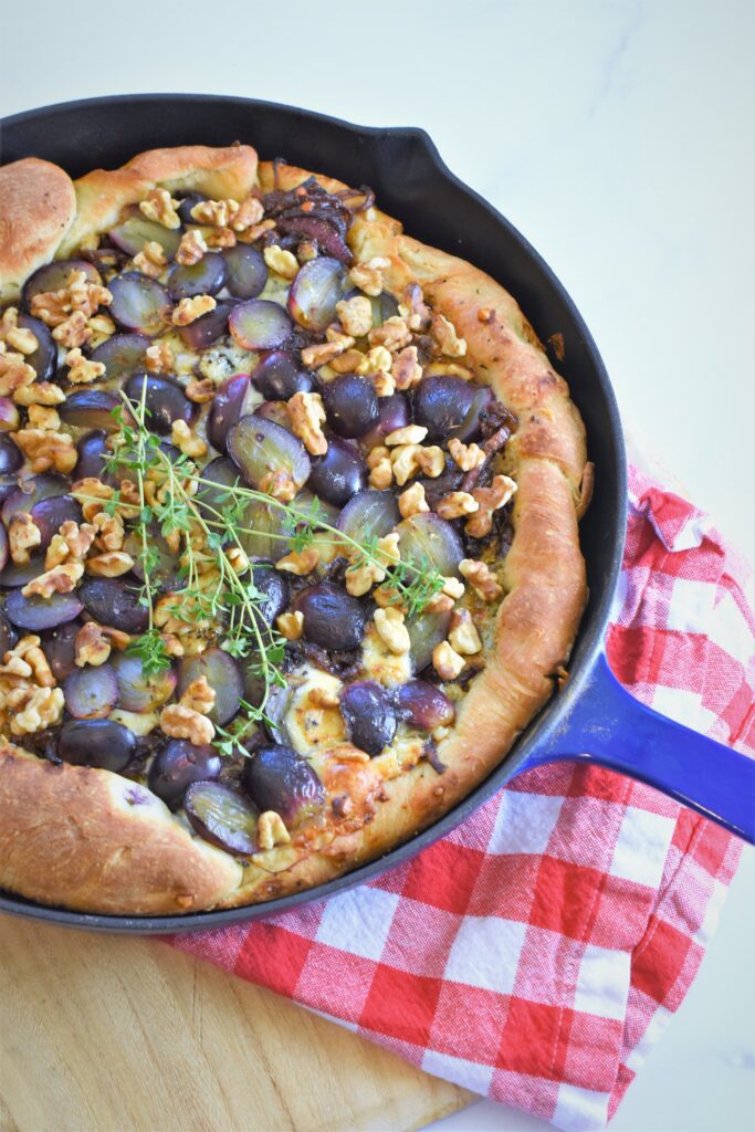 skillet black grape pizza with gorgonzola and walnuts in blue cast iron on red and white checkered napkin on wood cutting board 