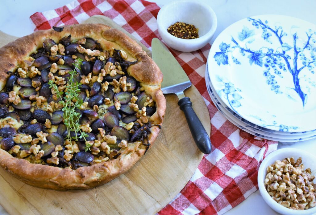 skillet black grape pizza pie with gorgonzola and walnuts with fresh thyme on wood cutting board red and white checkered napkin alongside a stack of blue and white grapevine themed plates with black marbled handled pie server and small white marble bowls of walnuts and chili pepper flakes 
