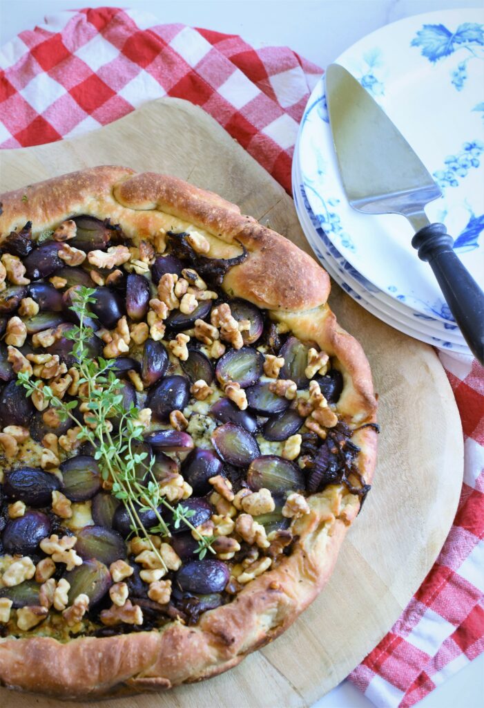skillet black grape pizza with gorgonzola and walnuts with fresh thyme on wood cutting board red and white checkered napkin with a stack of blue and white floral plates with black marble handled pie server