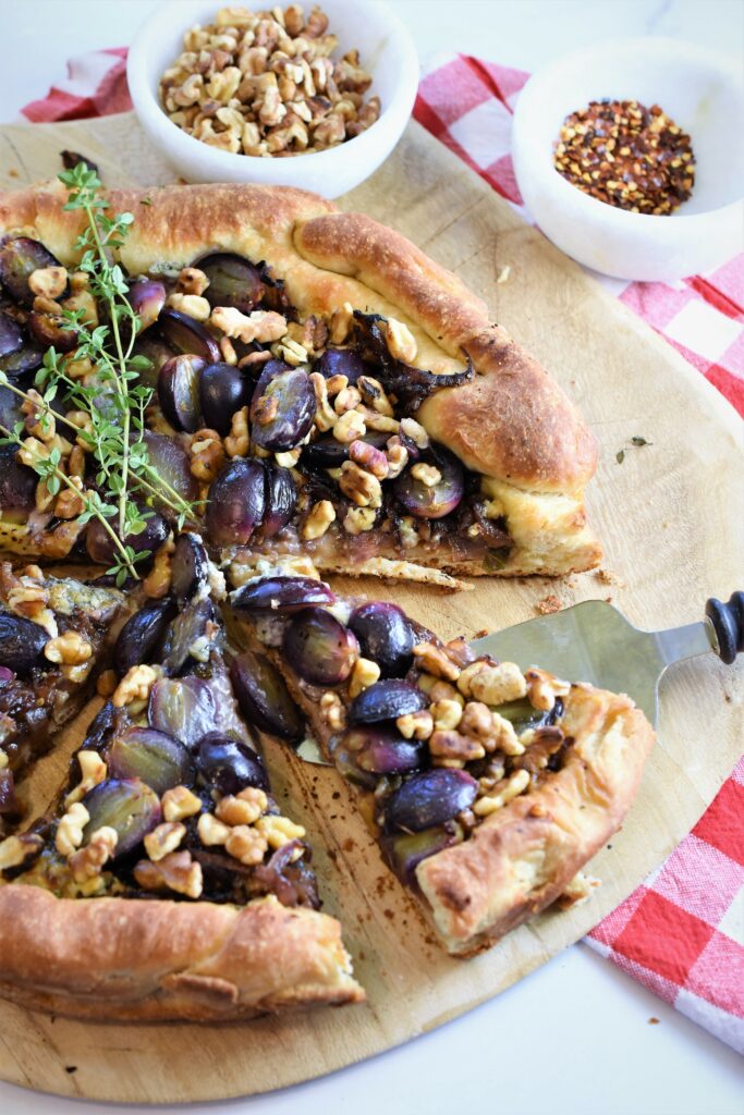 skillet black grape pizza with gorgonzola and walnuts with fresh thyme on wood cutting board with red and white checkered napkin along side small white marble bowls with walnuts in one and chili pepper flakes in another 