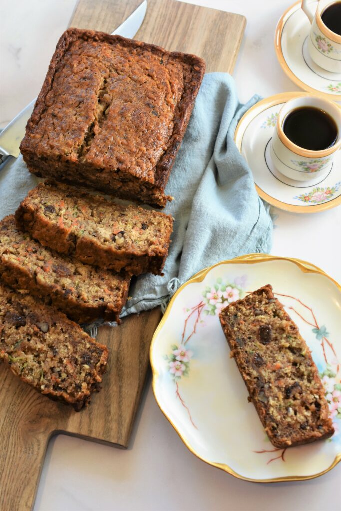 Banana Zucchini Carrot Bread with some sliced on cutting board with blue napkin along size antique floral plate with one slice next to two small cups of coffee in antique espresso cups and saucers 