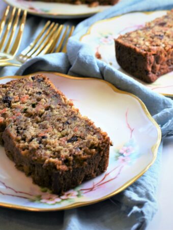 two floral and gold antique plates each with a slice of banana zucchini carrot bread along side gold forks and light blue napkin