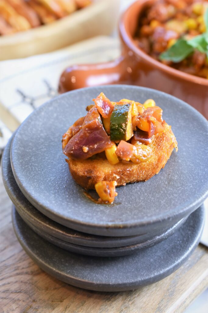 zucchini and corn capanata on toast on a stack of mini dark gray appetizer plates 