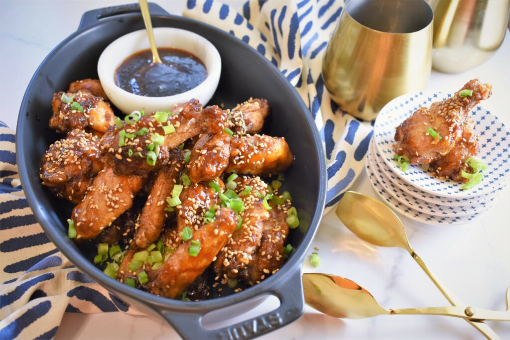 sesame apricot chicken wings served in oval black ceramic baking dish with small white marble dish with extra sauce on blue and white patterned cloth along with two brass stemless wine glasses off to the right with a stack of blue and white x pattern mini appetizer dishes with two wings displayed on top of those with brass serving tongs off to the side 