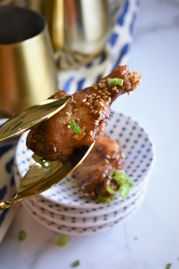 cup close image of brass serving tongs holding one roasted sticky Asian apricot chicken wing above appetizer plates 