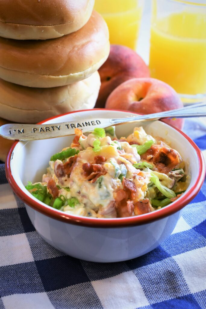 white and red enameled bowl fill with peach bacon & jalapeno schmear next to a stack of bagels with two peaches and two glasses of orange juice in the background