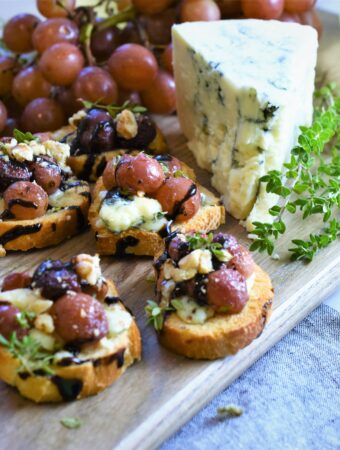 several crostini with roasted grapes on a cutting board with a hunk of blue cheese and red grapes
