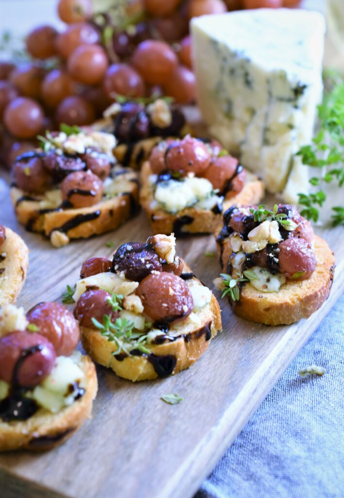 several servings of roasted grape bruschetta with blue cheese and walnuts with a large chunk of blue cheese to the right on a cutting board 