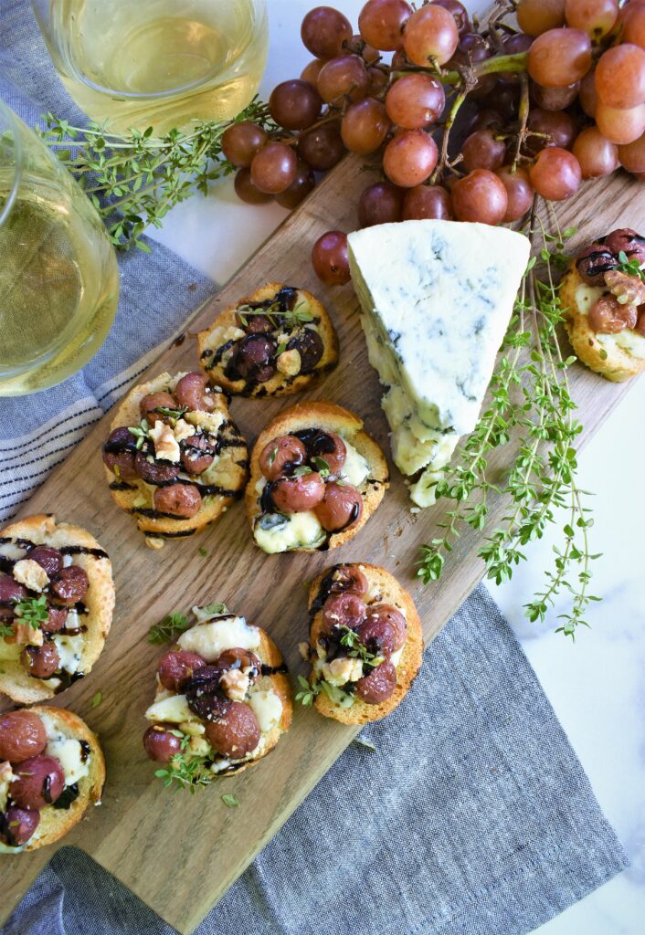 roasted grape bruschetta with blue cheese and walnuts on a cutting board with two glasses of wine off to the left