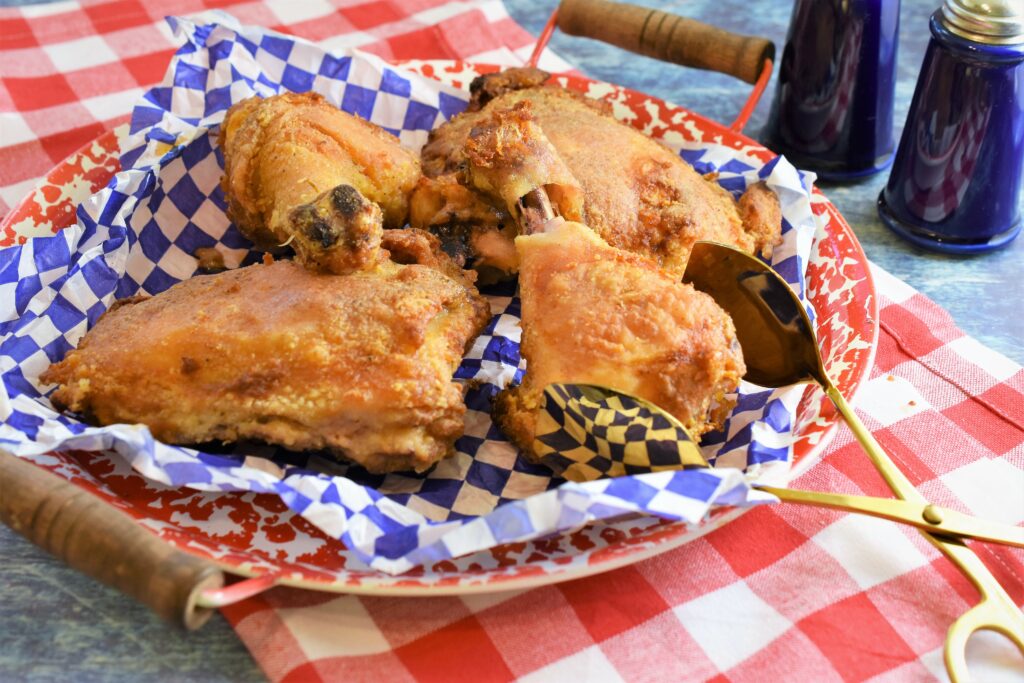 red and white splattered enamel platter with several pieces of oven fried chicken displayed on it with blue and white checked deli paper and gold serving tongs with cobalt blue salt and pepper shakers off to the side