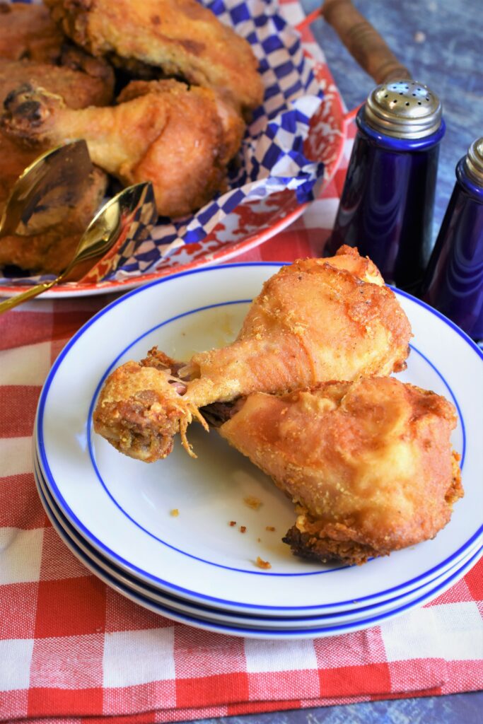 up close image of two oven fried chicken legs on a stack of white plates with a blue outer rim with a platter of chicken in the distance