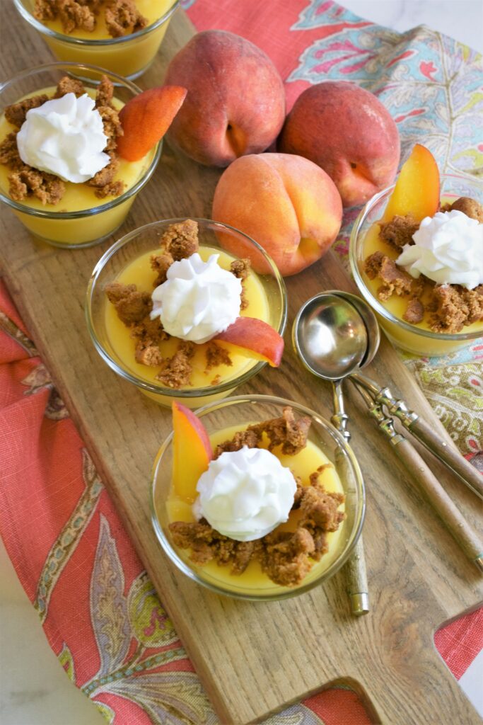 overhead image of vegan peaches and cream pudding in glass dessert bowls with wood handled teaspoons on cutting board and fresh peaches in the background