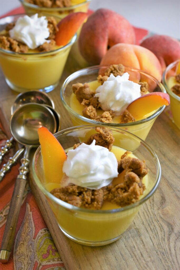 peach pudding garnished with whipped topping and crumbled cookies with a peach slice on cutting board with 3 stacked teaspoons off to the side on cutting board with fresh whole peaches in the background
