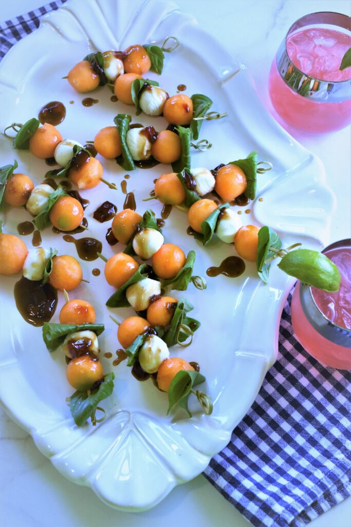 large oval white ceramic platter of Cantaloupe Caprese Skewers with Pepper Jelly Balsamic Glaze
with pink lemonade in a stemless wine glass off to the side with a blue and white checkered dish towel