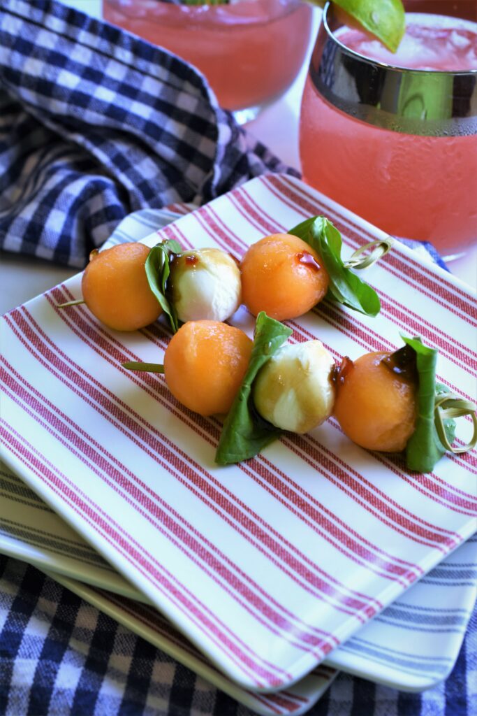 up close image of red and white striped appetizer plate with two mini skewered Cantaloupe Caprese Skewers with Pepper Jelly Balsamic Glaze