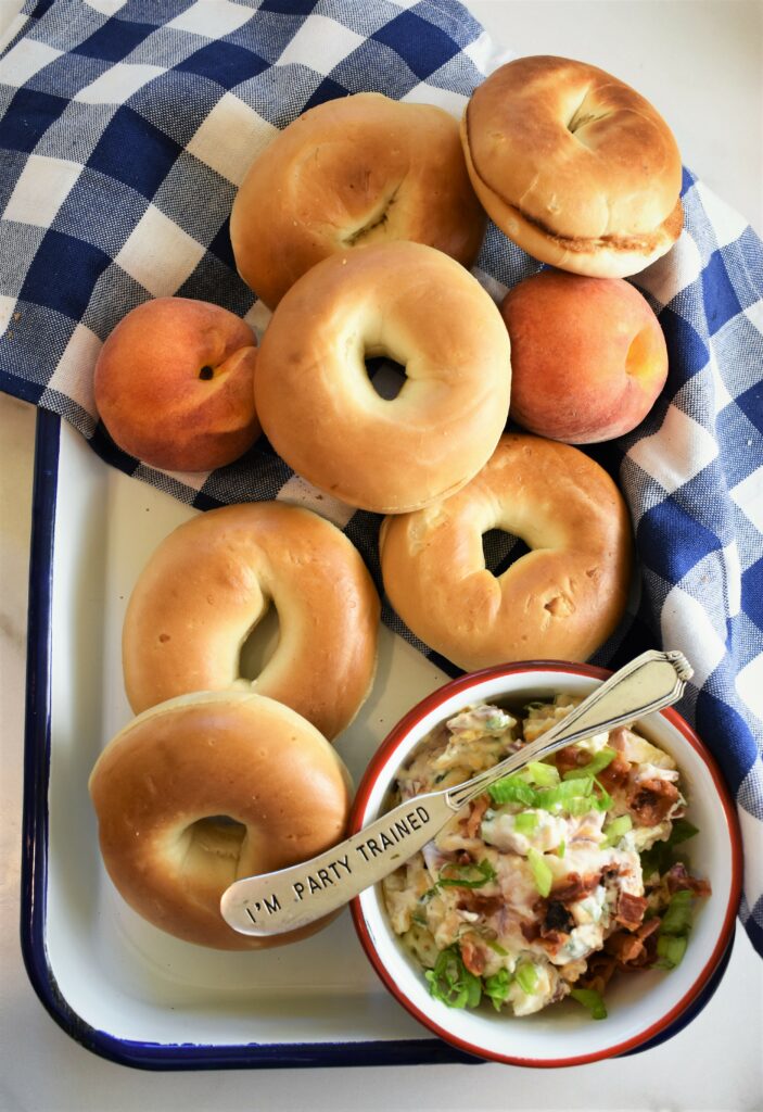 white and blue enameled tray with blue and white checkered napkin and a bowl of peach bacon & jalapeno schmear with spreading knife