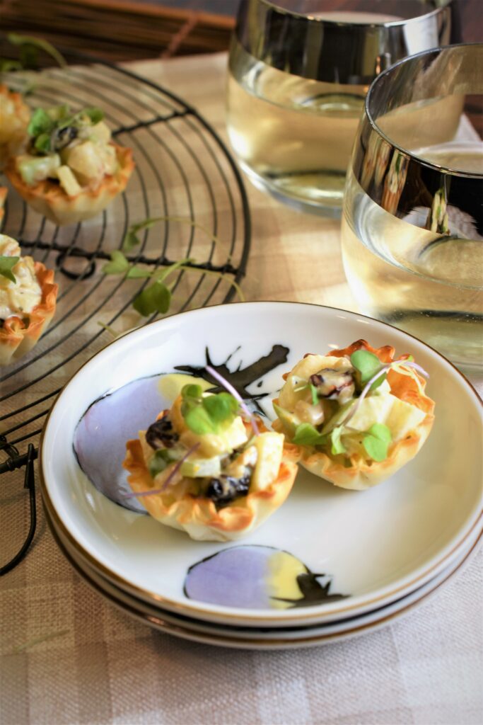 an appetizer plate with two mini waldorf salad in phyllo cups with a round wire cooling rack off to the side with more waldorf salad phyllo cups resting on it along with two glasses of white wine in the background