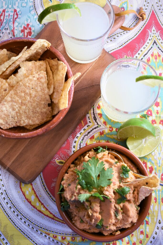 clay bowl of chipotle white bean dip with another bowl of tortilla chips and two margaritas off to the side