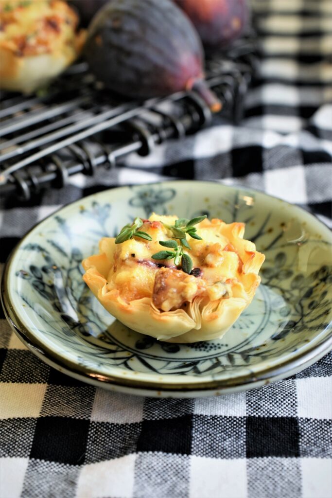 an up close image of a single fig, caramelized onion and blue cheese tart in a multi pattern mini ceramic dish with fresh figs in the background on a black and white gingham napkin