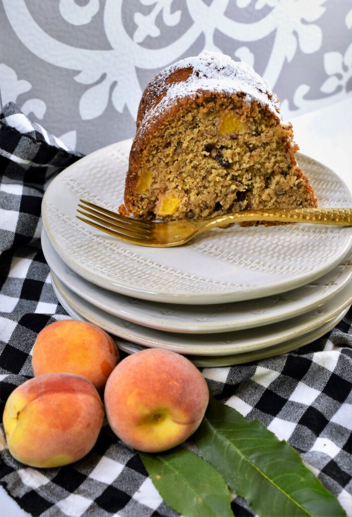 large slice of bundt cake on plate with peaches as garnish off to the side on a black and white gingham napkin