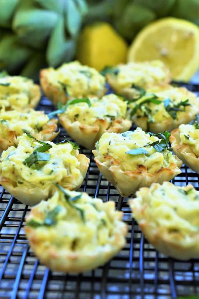 a dozen artichoke, ricotta & parmesan tarts displayed on a cooling rack with fresh artichokes and lemons in the background