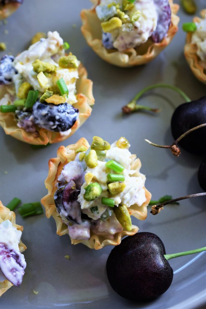 several chicken salad tarts with gorgonzola, pistachios & cherries displayed on a gray ceramic plate with fresh cherries on the stem off to the side 