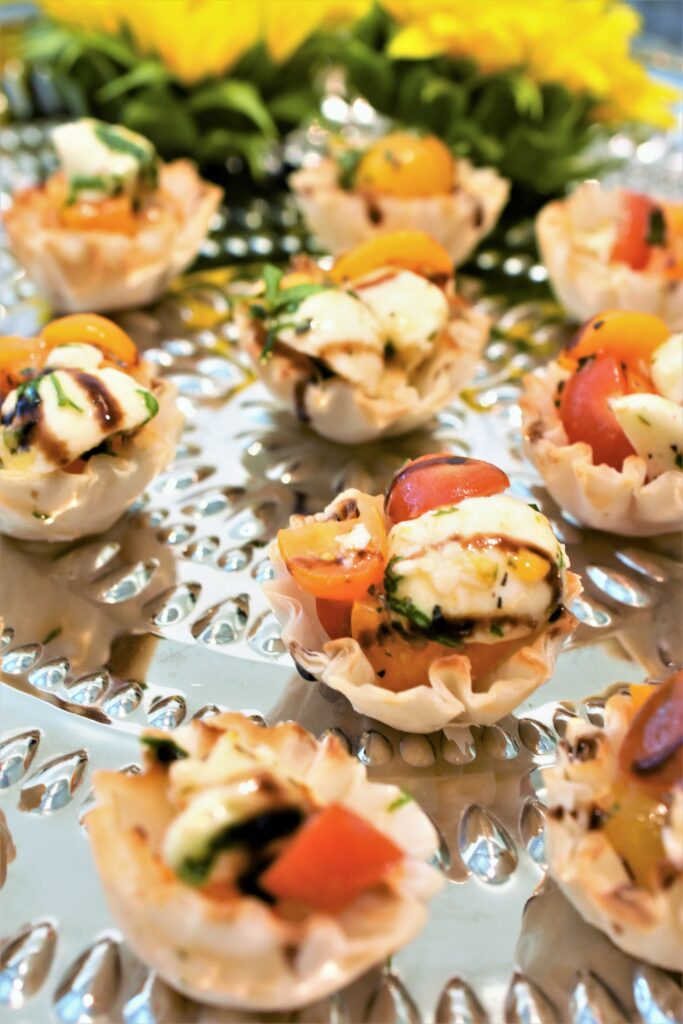 several caprese salad phyllo cups drizzled with balsamic glaze on a hammered silver tray garnished with a couple of sunflowers 