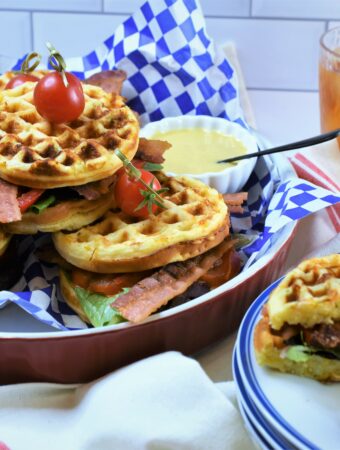 large full view of Jalapeno Cheddar Cornbread BLT Waffles in round platter and a serving cut in half on a stack of white plates with blue rim with two glasses of iced tea in the background