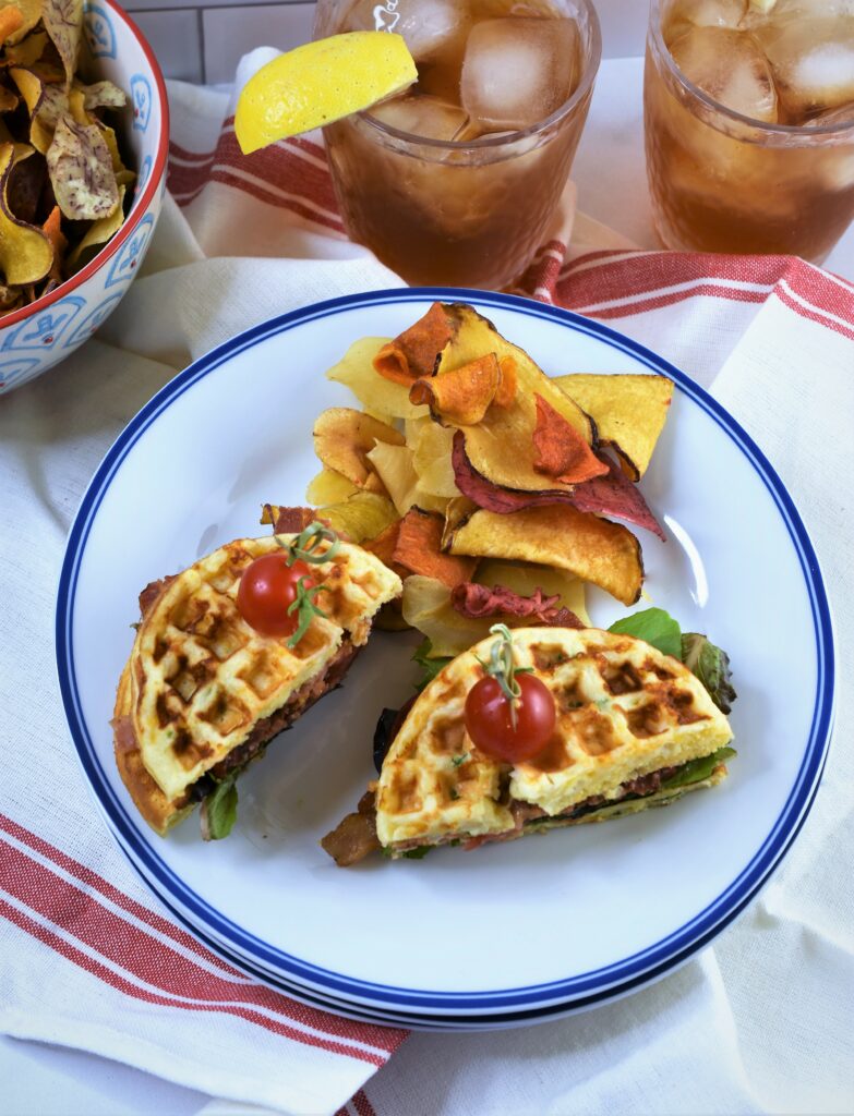 Jalapeno Cheddar Cornbread BLT Waffle cut in tow served on a blue rimmed white bistro cafe plate alongside crispy vegetable chips on white towel with a few red stripes with two iced teas in the background