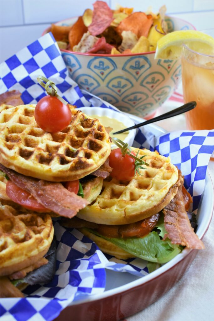 up close photo of Jalapeno Cheddar Cornbread BLT Waffles in red ceramic quiche dish lined with red and white checkered paper garnish with cherry tomatoes on a pick with a small bowl of honey Dijon mustard mayo in a rippled white round dish with ceramic bowl in the background with vegetable chips with an iced tea to the righ