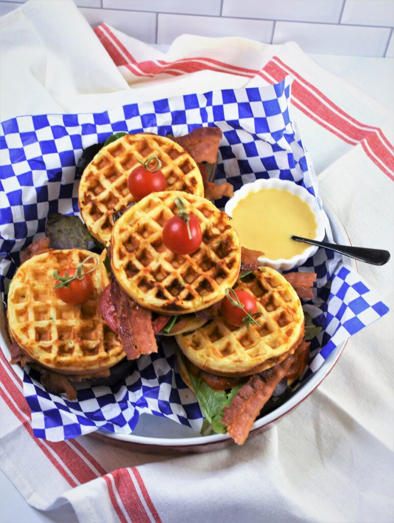 overhead view of breakfast sandwiches in round red quiche plate lined with blue and white checked deli sandwich paper with small bowl of honey Dijon mustard mayo spread on white towel with red striped border on counter top with subway tile back splash