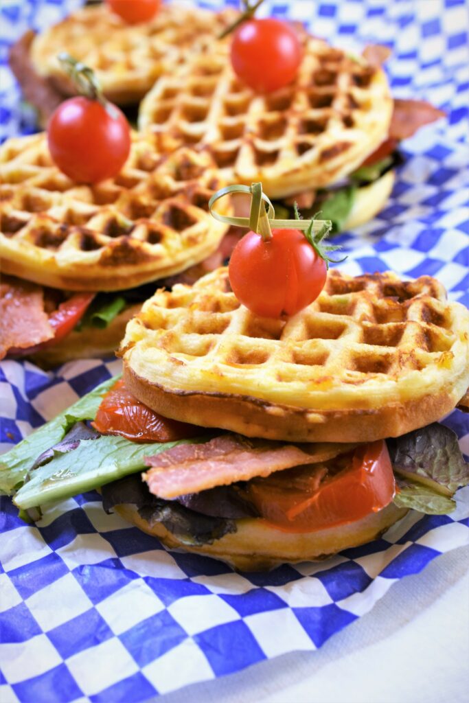 up close view of Jalapeno Cheddar Cornbread BLT Waffles of four of them staggered on blue and white checkered paper garnished with cherry tomato sandwich picks