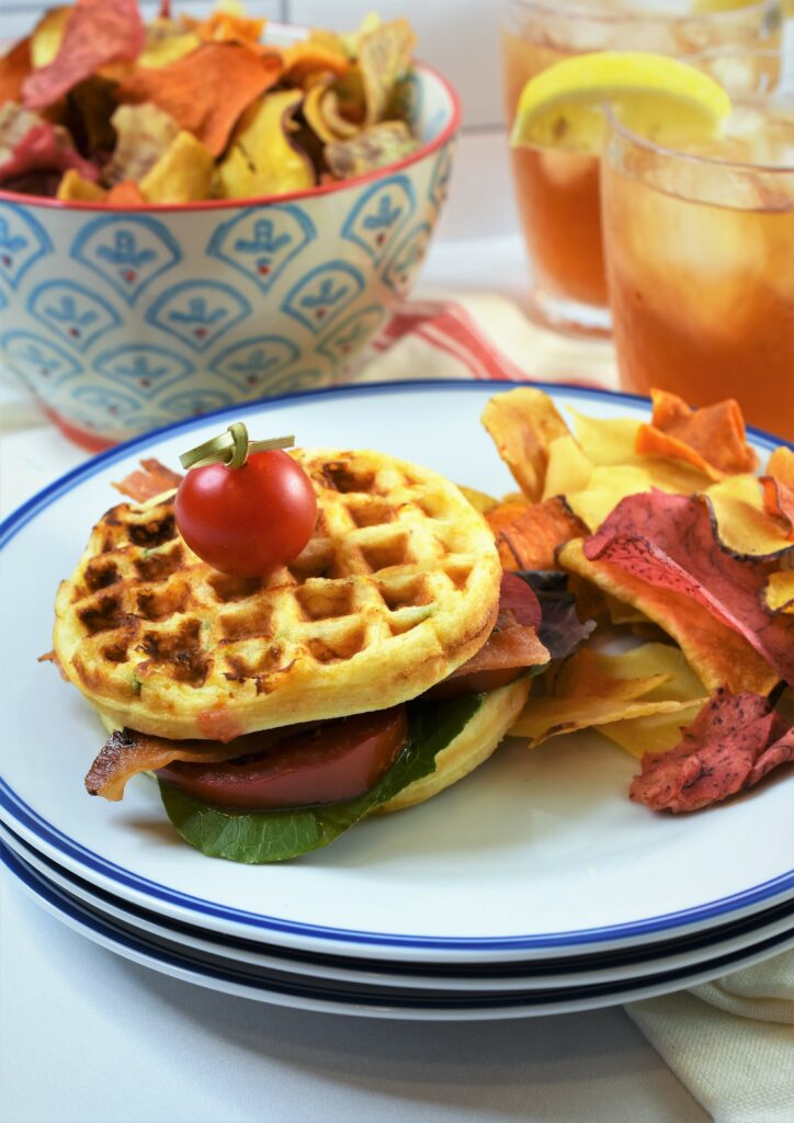 one plate with sandwich garnished with a cherry tomato pick with vegetable chips with a bowl of vegetable chips in the background with two iced teas in the background