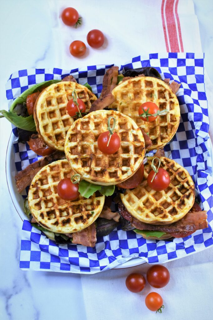 overhead photo of BLT waffle sandwiches in blue and white checkered paper lined round platter garnished with cherry tomatoes on white towel with a couple of red stripes in it