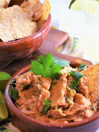 clay bowl of creamy chipotle white bean dip with tortilla chips in another clay bowl off to the side on a small wood cutting board