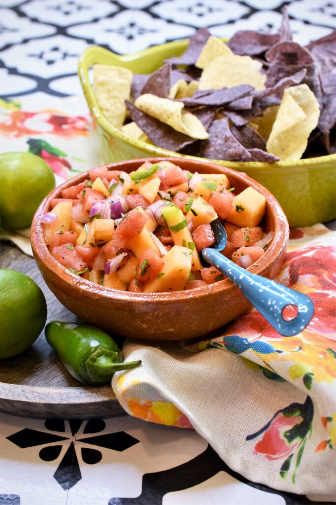 clay mexican style bowl of watermelon and cantaloupe salsa along side a lime colored ceramic handled bowl of blue and white tortilla chips on a rose floral napkin on a wood platter