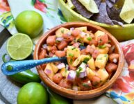 above image shot of clay mexican style bowl of watermelon and cantaloupe salsa along side a lime colored ceramic handled bowl of blue and white tortilla chips on a rose floral napkin on a wood platter