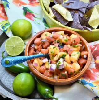 above image shot of clay mexican style bowl of watermelon and cantaloupe salsa along side a lime colored ceramic handled bowl of blue and white tortilla chips on a rose floral napkin on a wood platter
