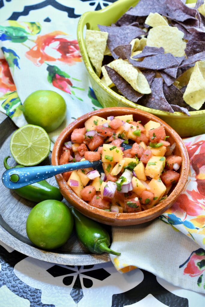 above image shot of clay mexican style bowl of watermelon and cantaloupe salsa along side a lime colored ceramic handled bowl of blue and white tortilla chips on a rose floral napkin on a wood platter