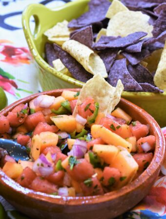 clay mexican style bowl of watermelon and cantaloupe salsa along side a lime colored ceramic handled bowl of blue and white tortilla chips on a rose floral napkin on a wood platter