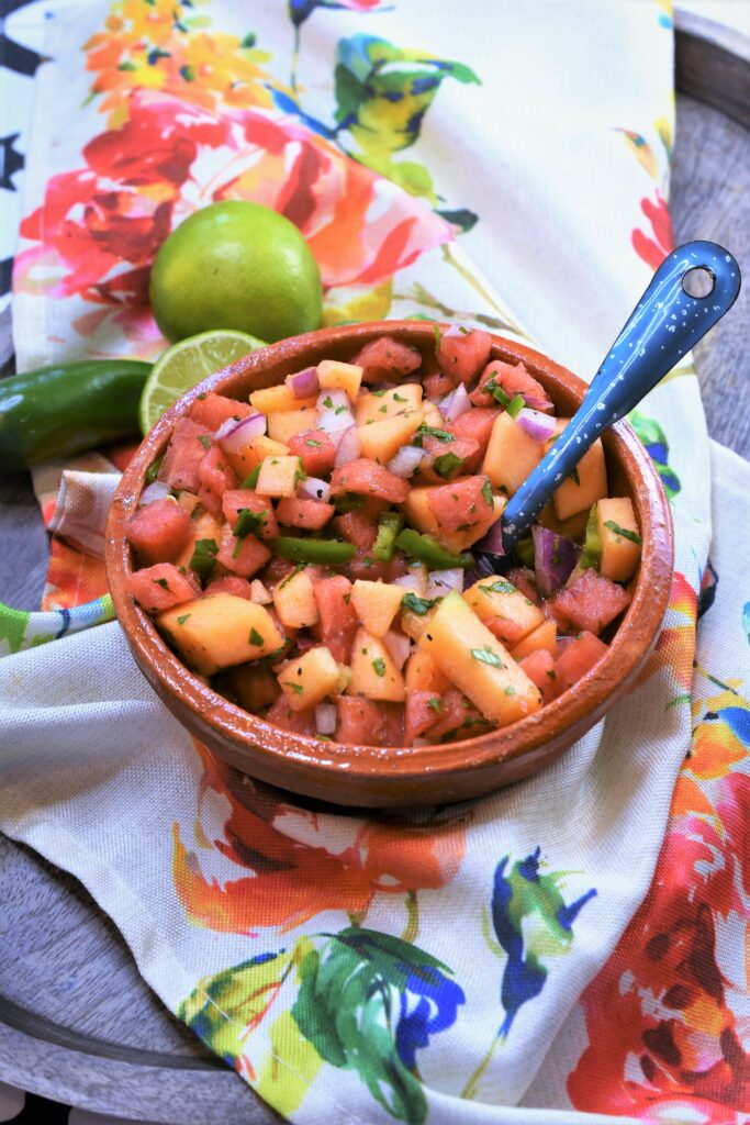 clay bowl of fruit relish on bright rose floral napkin on wood platter with fresh limes and jalapeno in the background