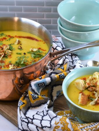 copper pot of cauliflower soup with soup bowls ready to serve on wood board