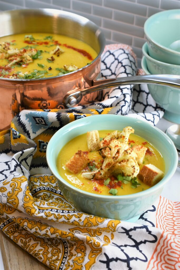 cutting board with bowl of soup and copper pot of soup placed on it with colorful patterned cloth napkin