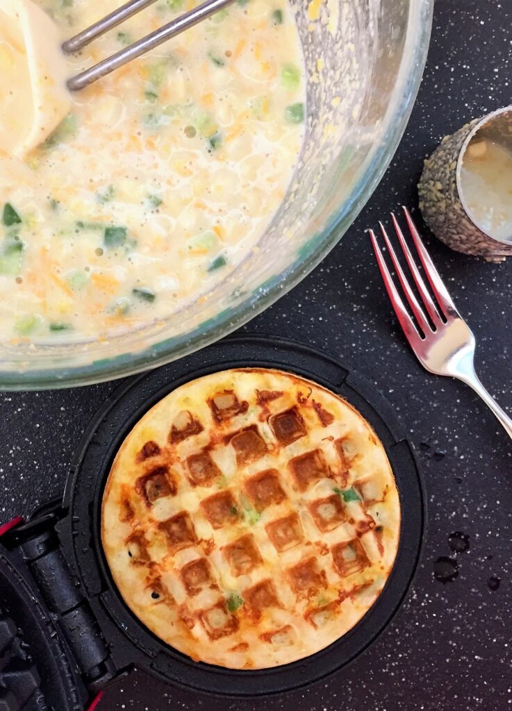 overhead view of bowl of batter with for and measuring cup off to the side with fresh cooked waffle in maker