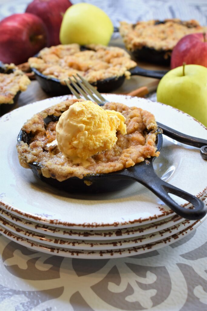 image of cast iron apple crisp on a stack of dessert plates with more in the background along with whole apples