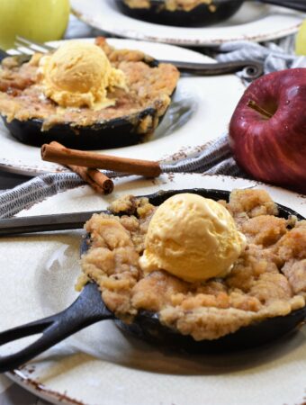 small cast iron skillets of apple crisp on rustic white plates on gray and white spanish tile countertop with assorted colored apples and cinnamon sticks with gray and white patterned kitchen towel