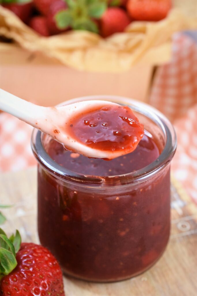 up close image of small white spoon with red pepper strawberry jam above a glass pot of jam
